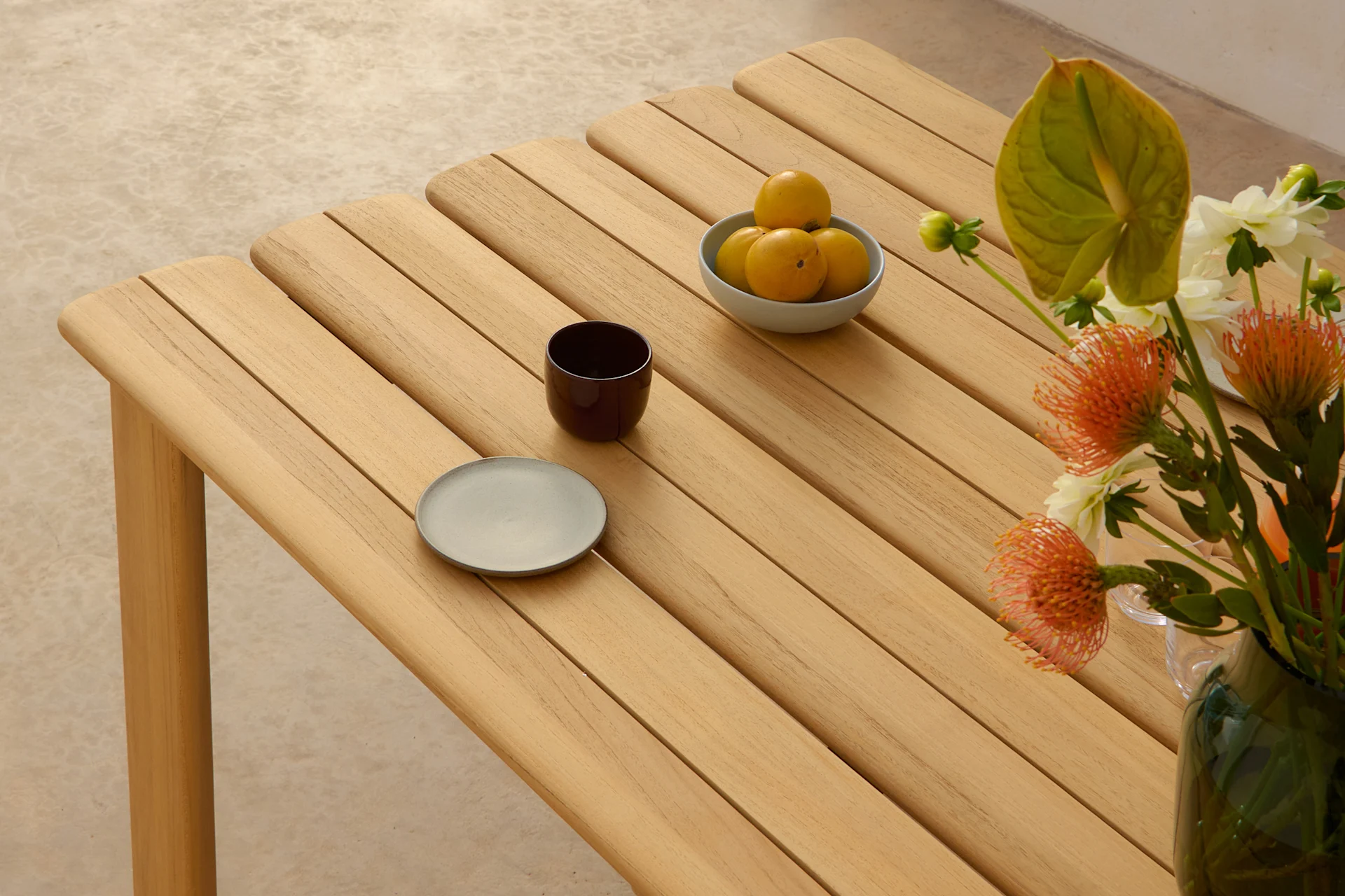 Et lyst trebord med lameller, med en skål gule frukter, en mørk kopp, en grå tallerken og en vase med oransje og hvite blomster.