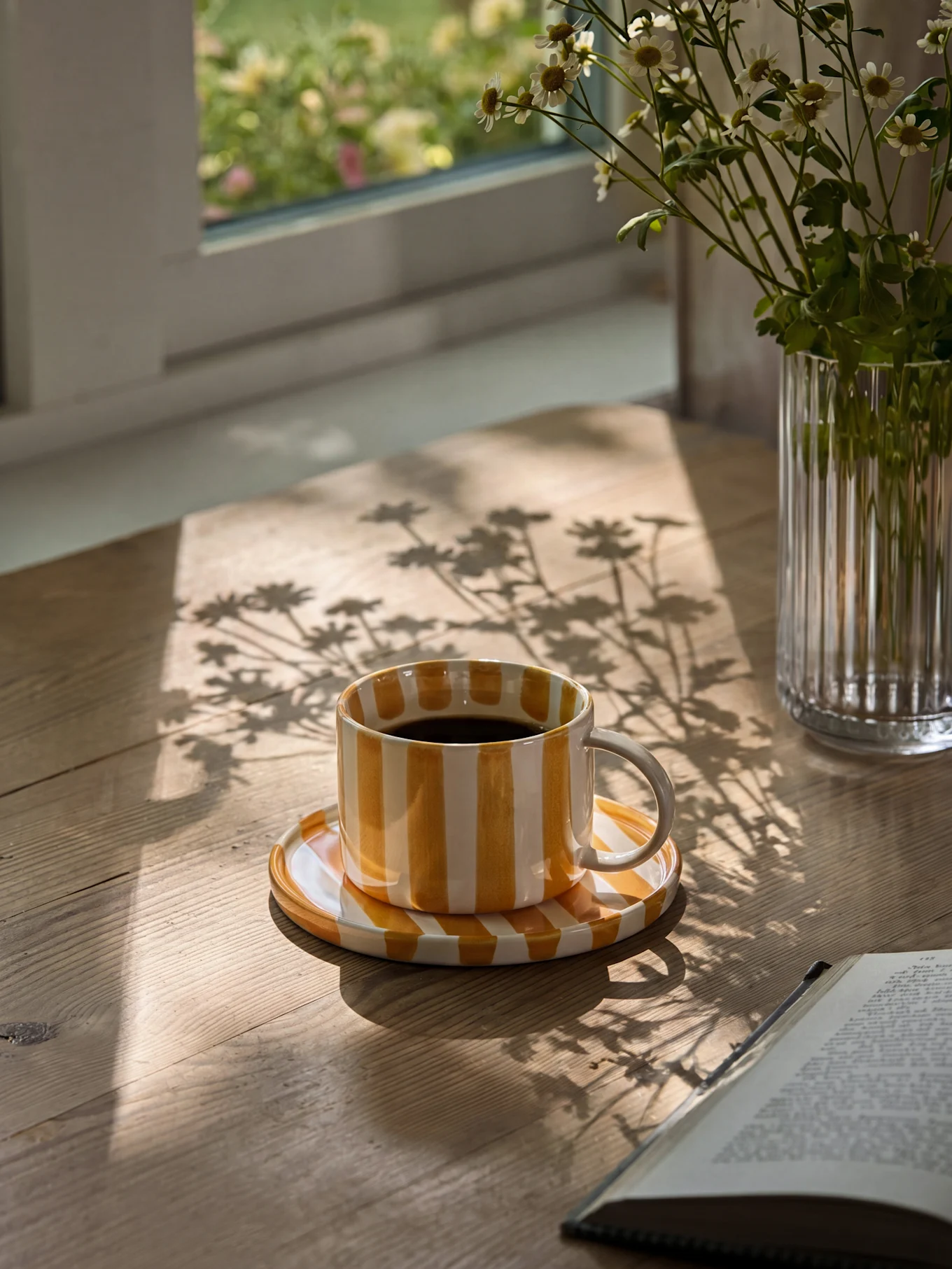 Gul og hvitstripet kaffekopp med svart kaffe på et trebord med solskygger og en vase med tusenfryd.