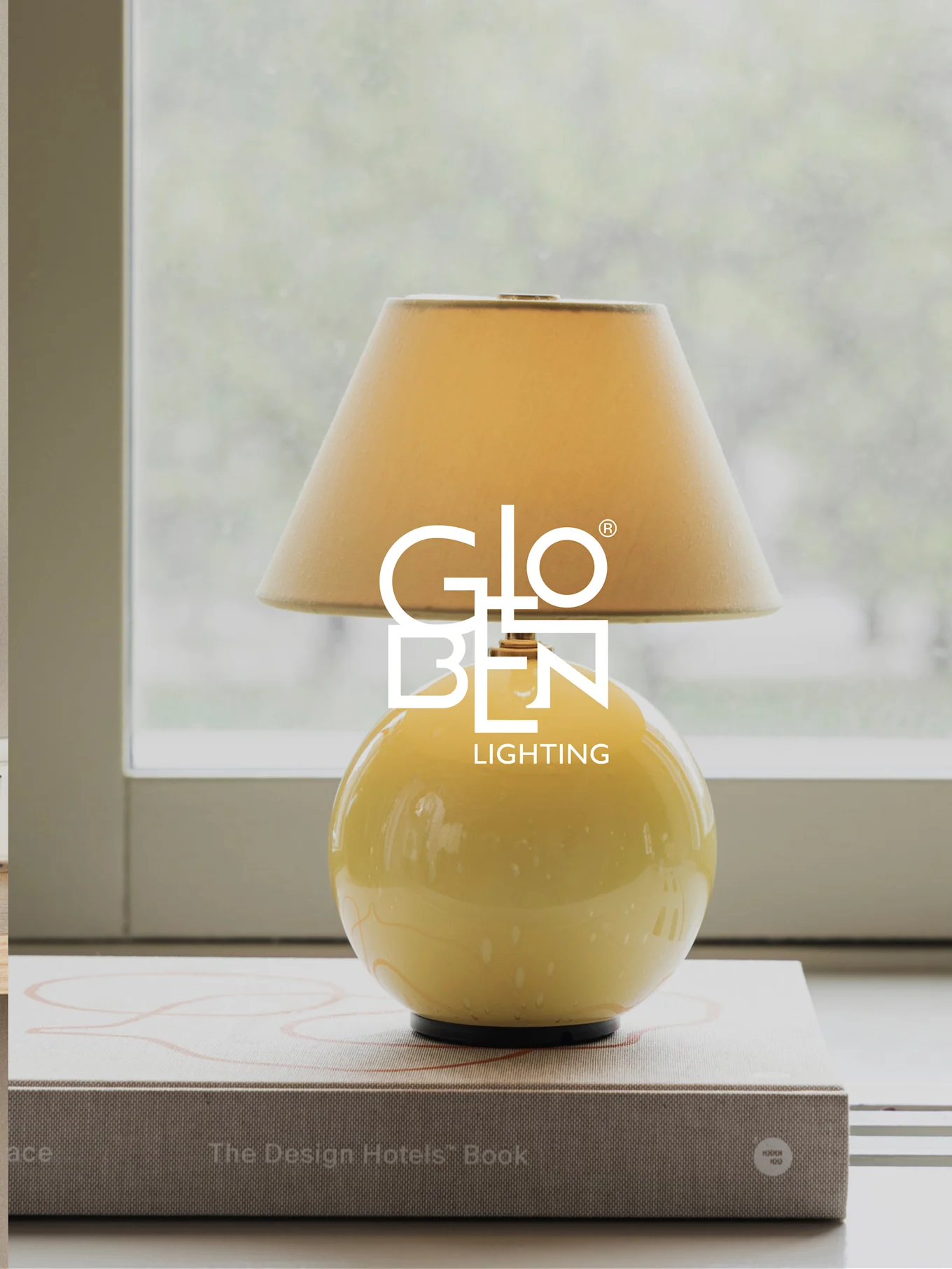 Gul bordlampe med beige skjerm, står på bøker ved et vindu.
