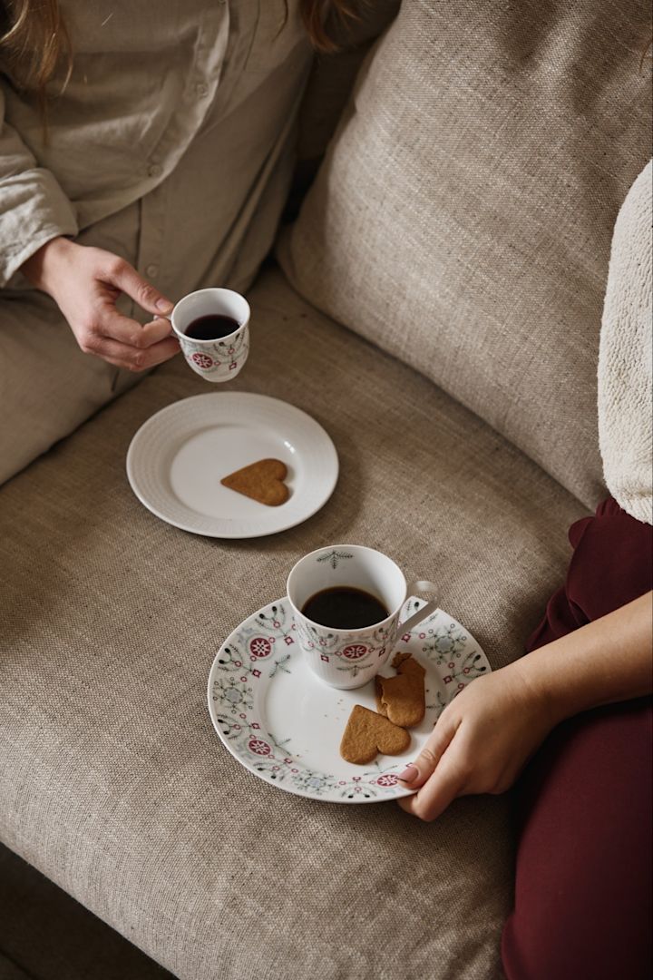 Et par sitter i en sofa med Swedish Grace Winter-kolleksjonen. Her ser du både en vanlig kopp og en gløggkopp, samt tallerkener med pepperkaker.