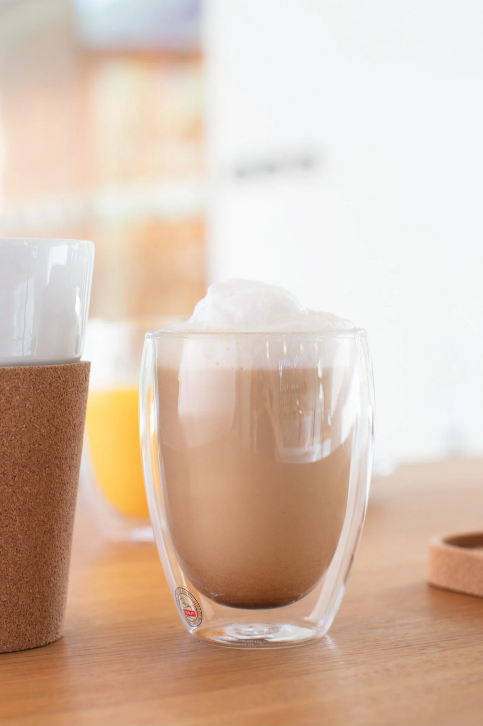 Skummende latte i et dobbeltvegget klart glasskrus på et trebord, med et annet krus med korkmansjett til venstre og en oransje drikke i bakgrunnen.