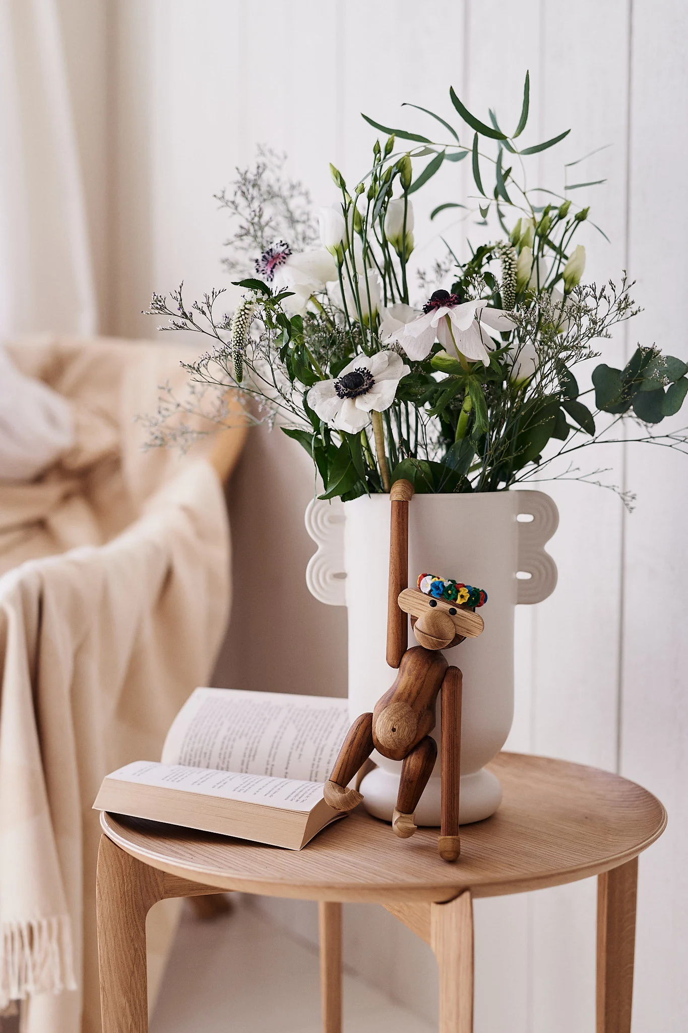 En treapeleke henger fra en hvit vase fylt med hvite blomster ved siden av en åpen bok på et trebord.