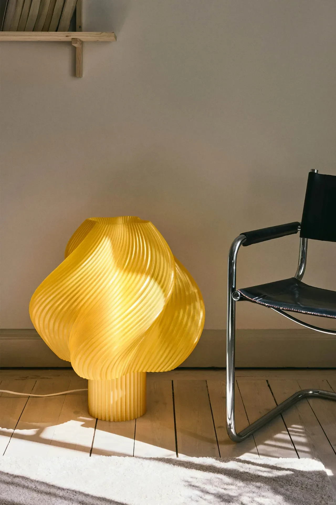 En lys gul, skulpturell bordlampe med riflet, bølget design lyser på et tregulv ved siden av en sort skinn- og kromstol.