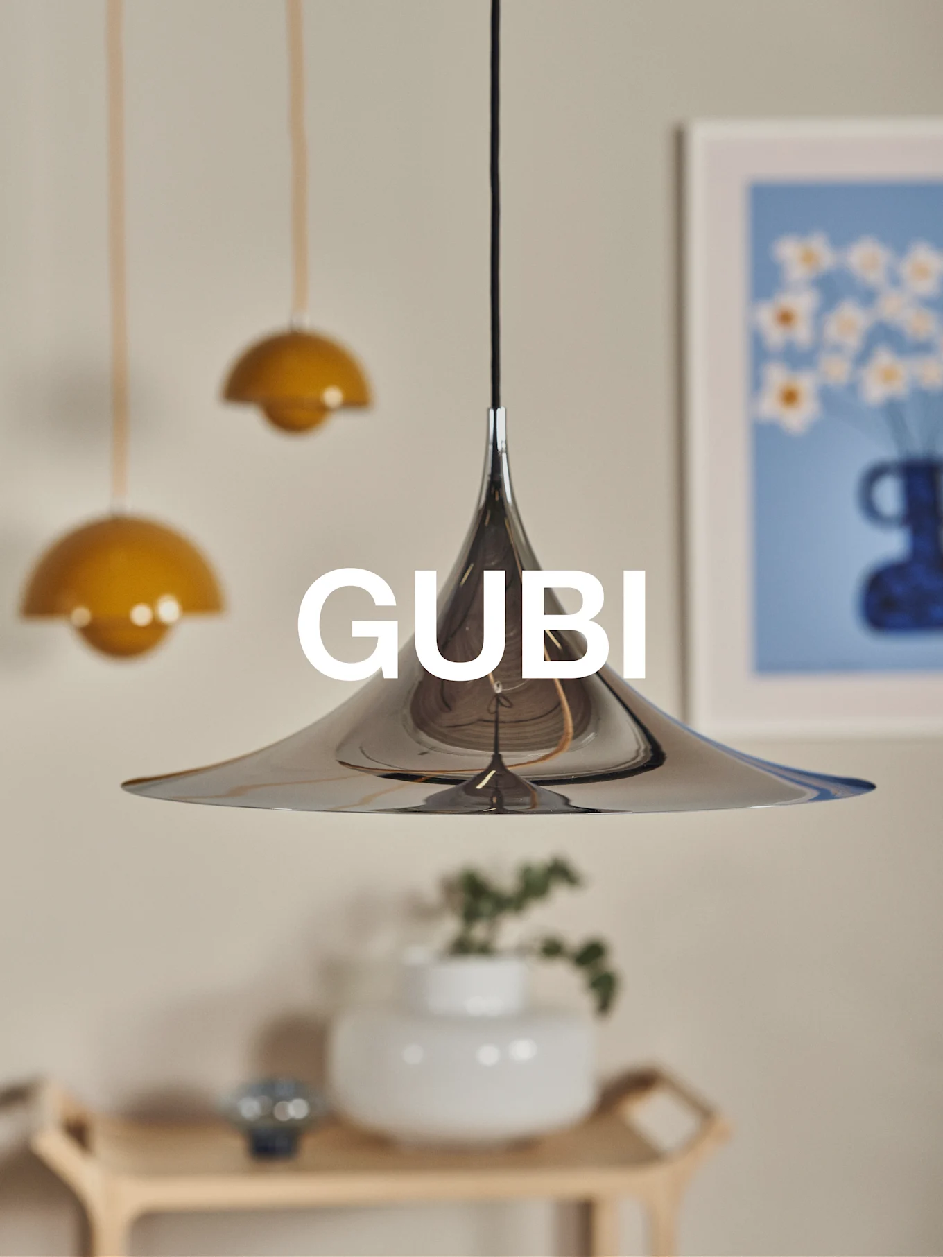 En polert konisk pendellampe i krom med GUBI-logoen, mot en uskarp bakgrunn med andre lamper og kunst.