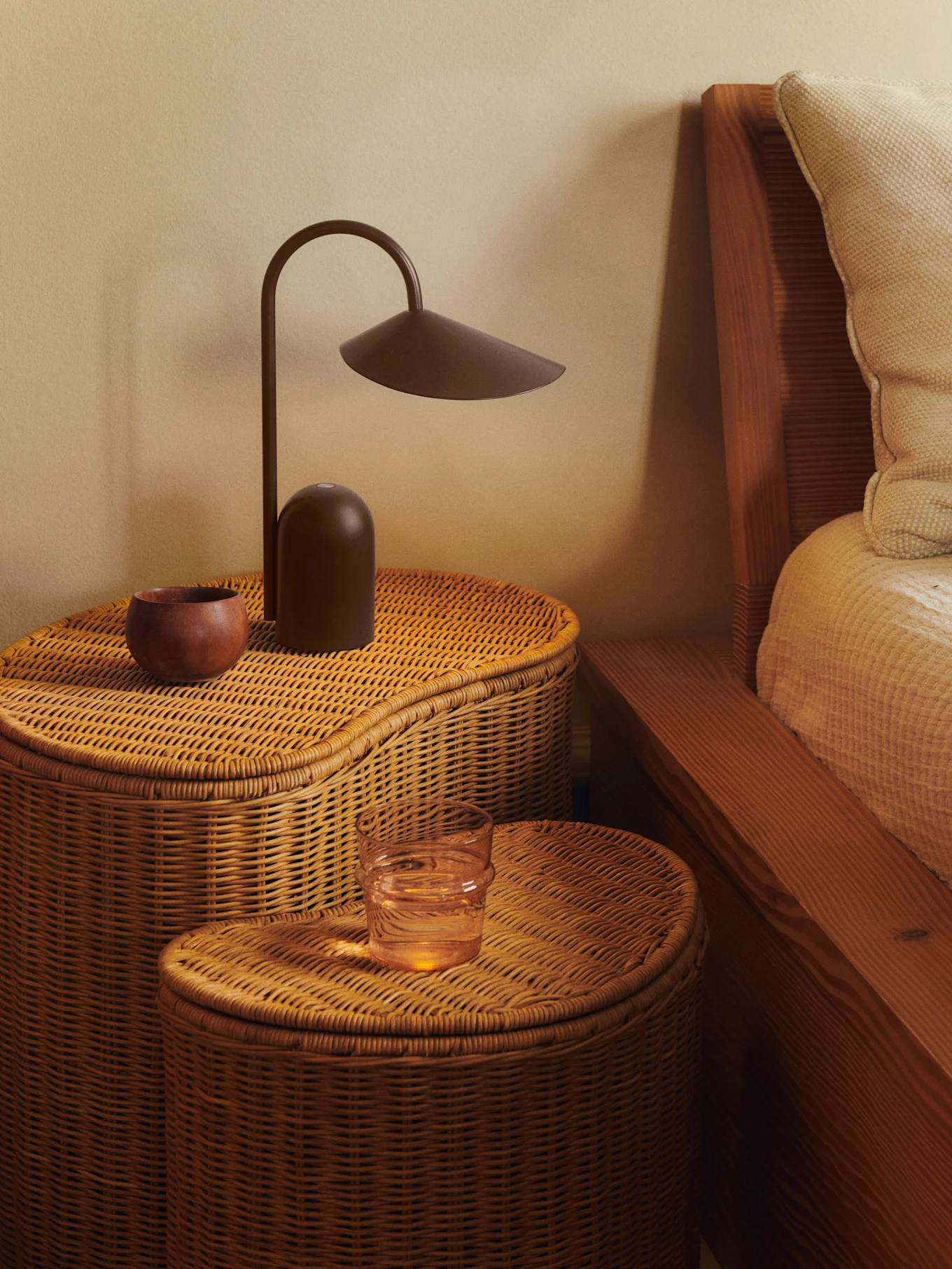 To flettede nattbord i rotting med en mørkebrun lampe, en treskål og et glass vann.