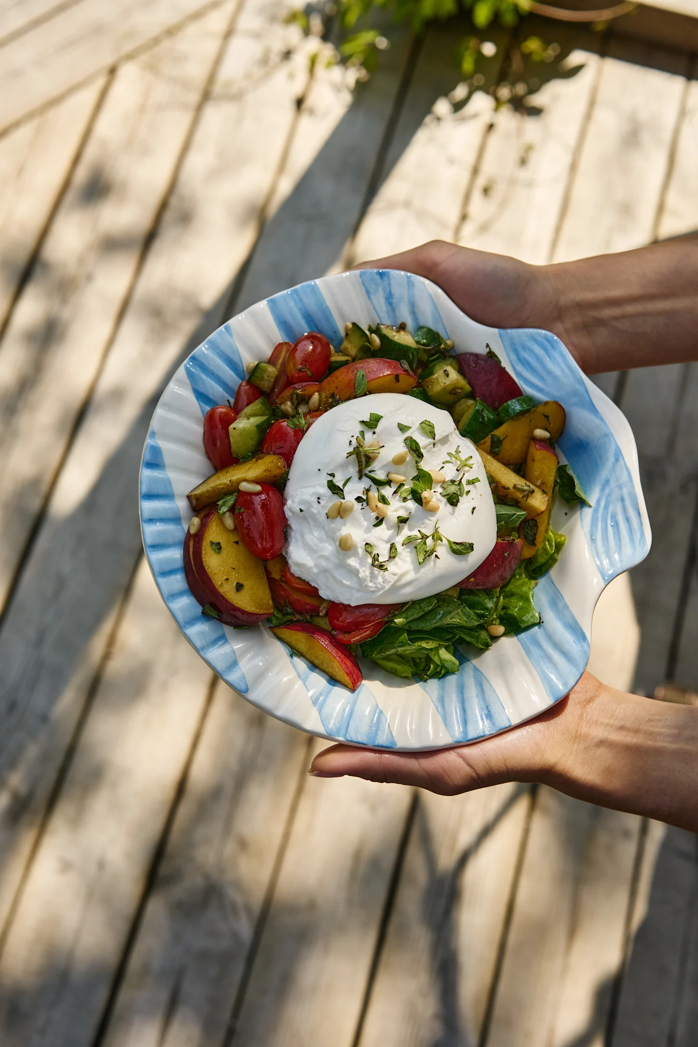 Hender holder en burrata-salat med tomater, fersken, agurk og grønt i en stripete skjellformet bolle.