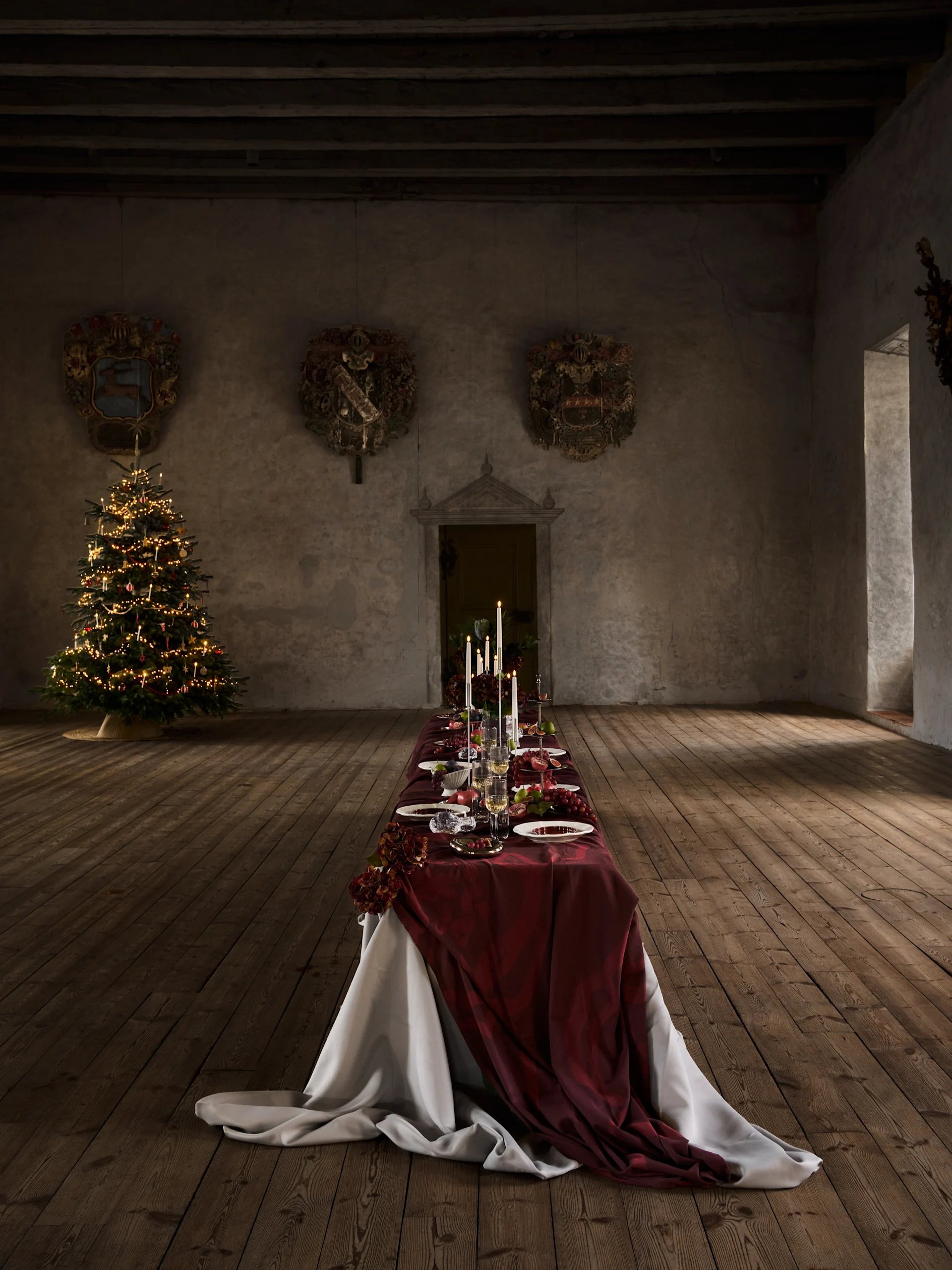 Langt julemiddagsbord med rød duk, levende lys, frukt og servise, ved siden av et tent juletre i en gammel sal.
