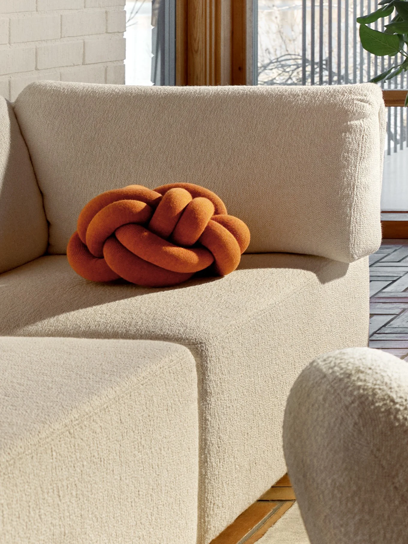 En oransje knutepute på en beige teksturert sofa, med en hvit murvegg og et vindu i bakgrunnen.