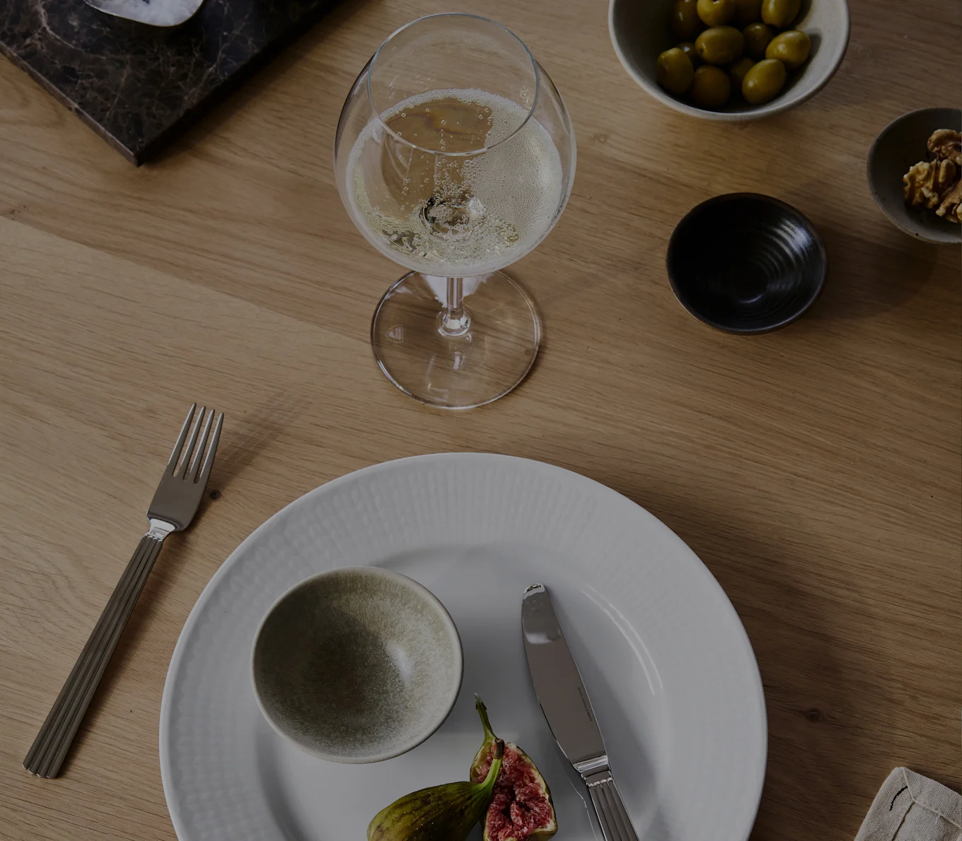 Et glass champagne, hvit tallerken med bolle og fiken, gaffel, kniv, oliven og valnøtter på et trebord.