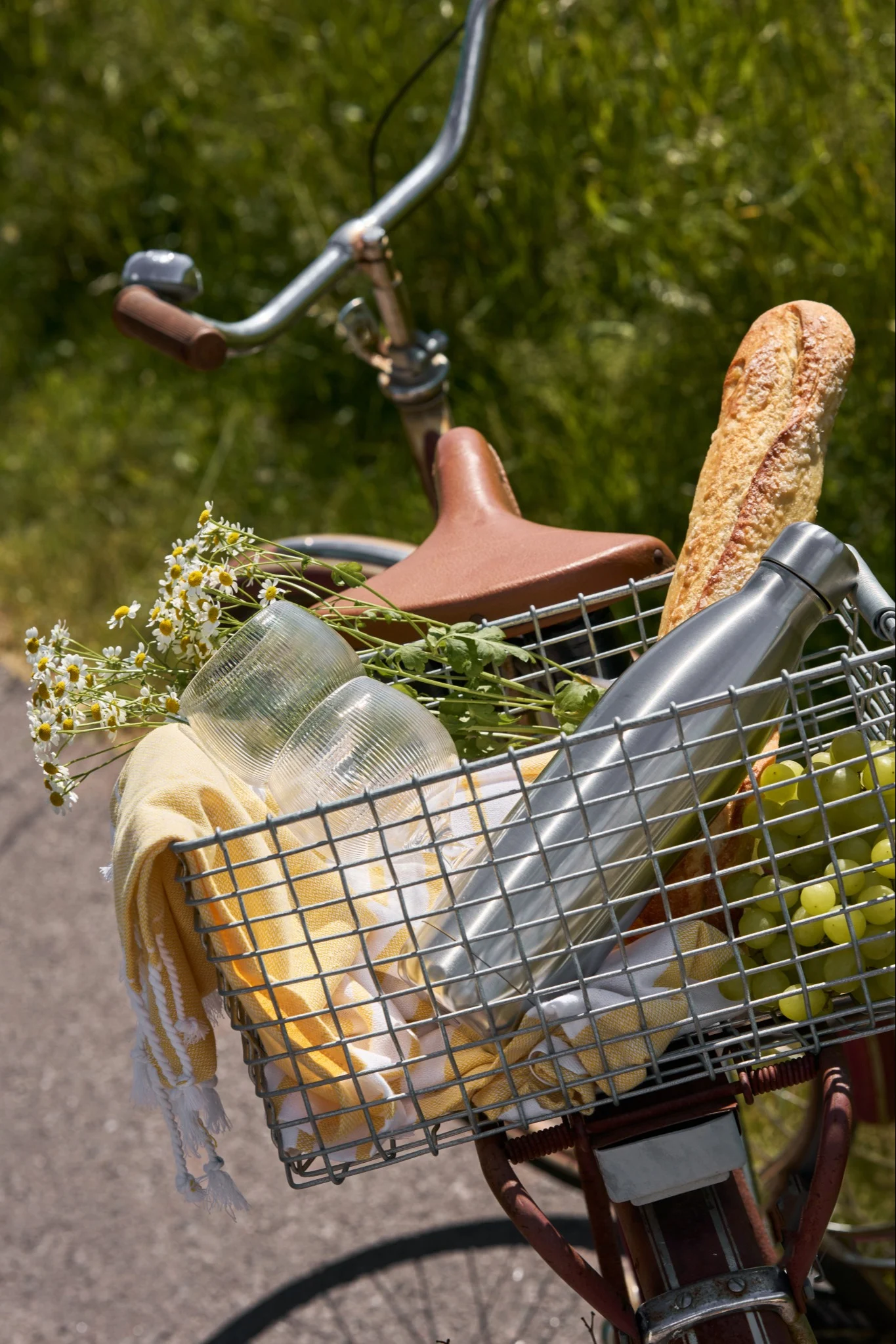 Sykkel med en frontkurv fylt med pikniksaker: brød, grønne druer, en sølv vannflaske, kopper, et gult håndkle og blomster.