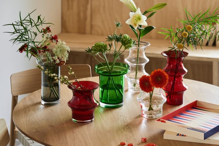 En samling røde, grønne, grå og klare vaser står på et bord med et rødt og blått backgammonsett.