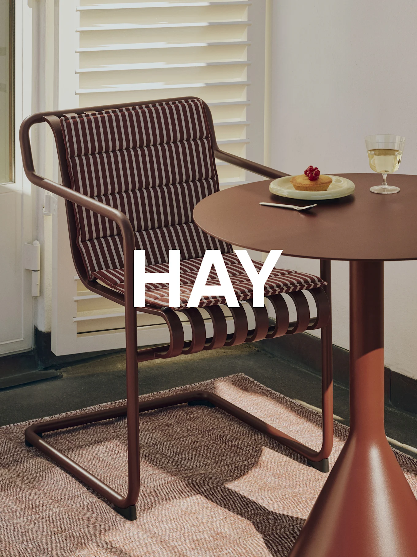 Uteplass med brun metallstol, stripete pute, matchende bord, terte, vinglass og 'HAY'-merkevare.