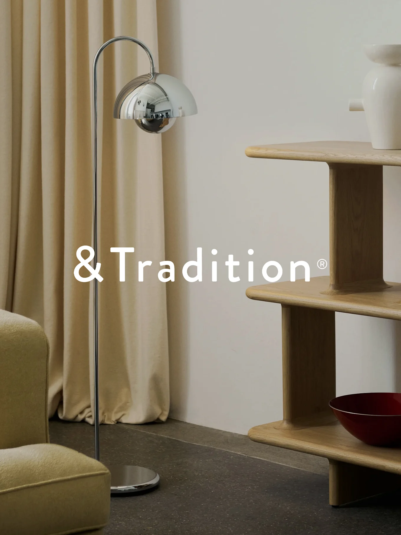 Et moderne interiør med en krom gulvlampe, en hyllereol i lyst tre med flere nivåer og en lenestol. &Tradition®-logo.