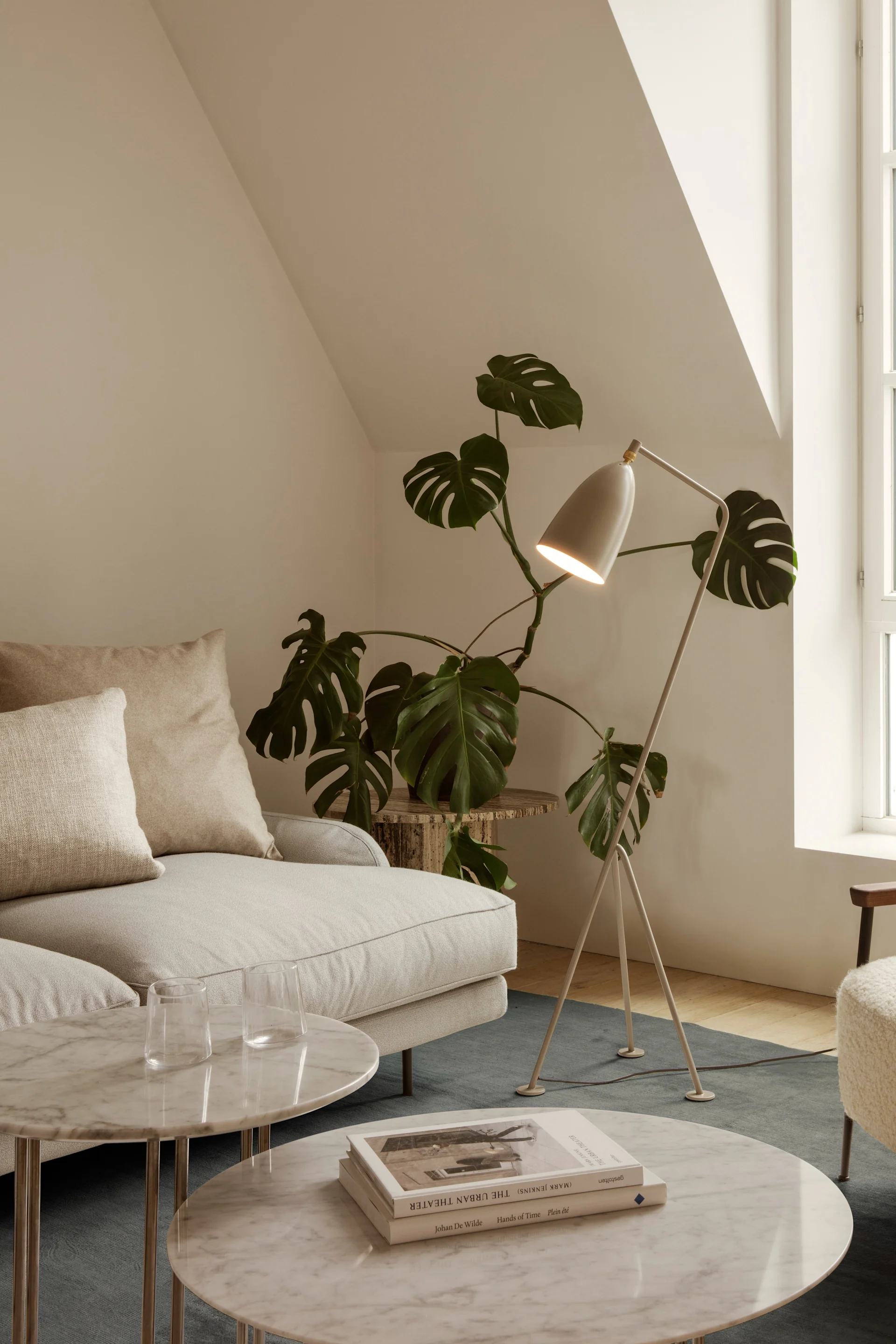 En lys stue med en kremfarget sofa, marmorbord, en gulvlampe, en plante og en myk lenestol ved et buet vindu.