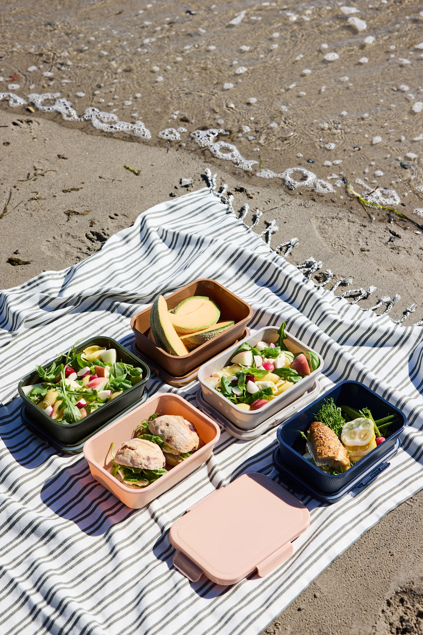 Fem åpne, fargerike matbokser med salater, smørbrød og melon, pluss en lukket rosa boks, på et stripet strandteppe.