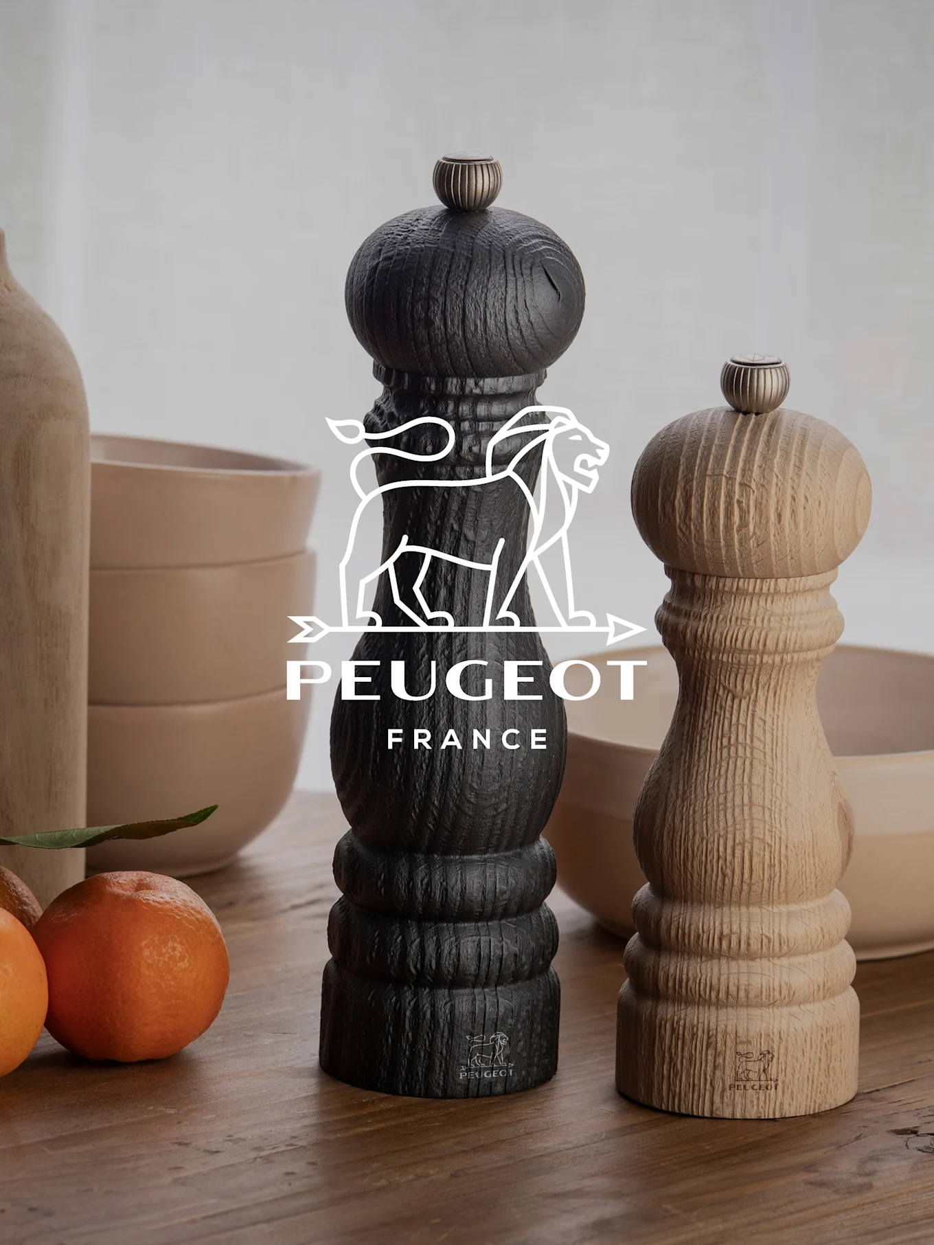 To Peugeot pepperkverner, en svart og en i naturtre, på et trebord med mandariner og skåler.