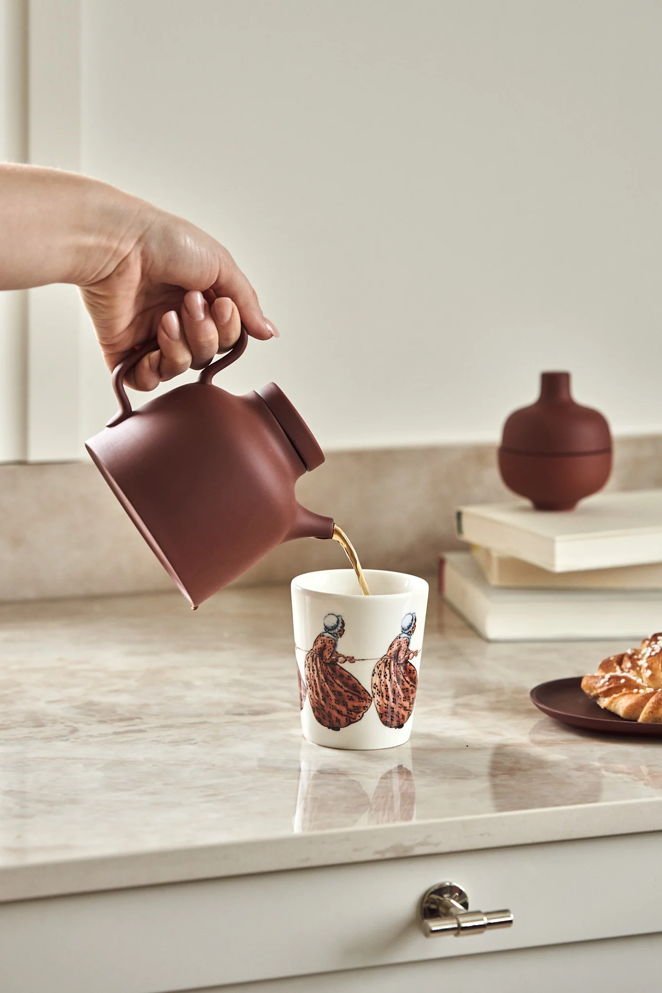 Krus med Elsa Beskow-motivet Tant Brun på kjøkkenbenk sammen med kanna Sand i terrakotta-rødt.