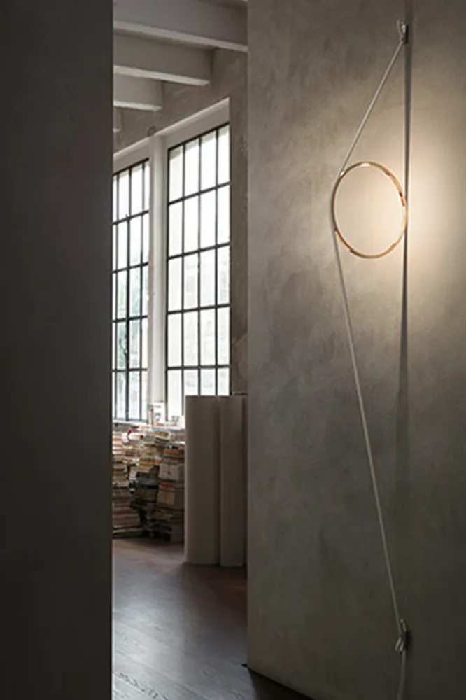 En moderne sirkulær lysarmatur hengt opp med snorer på en betongvegg, med store vinduer og bøker i bakgrunnen.