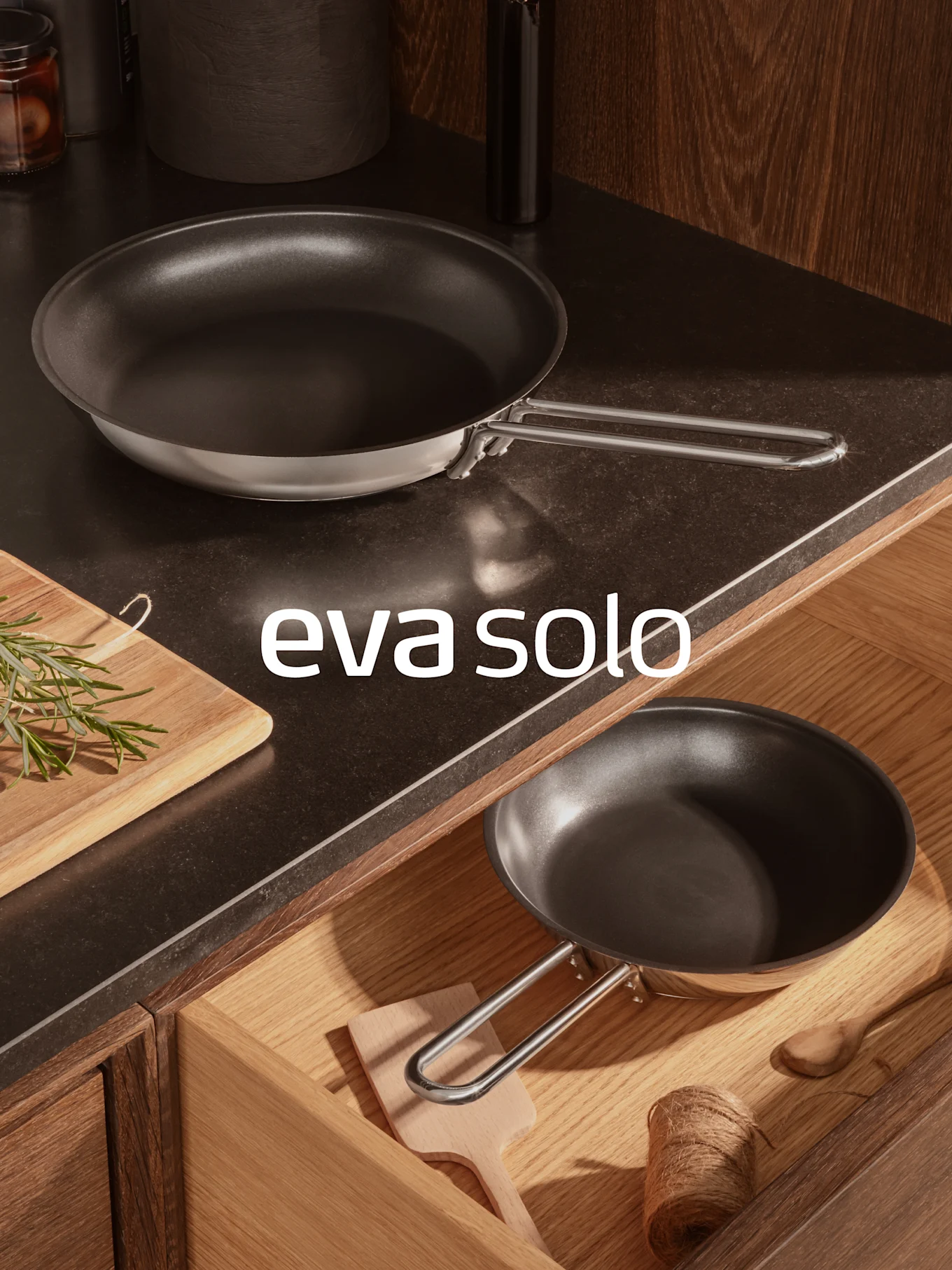 To sorte stekepanner, en på en mørk kjøkkenbenk med rosmarin, en i en åpen treskuff. 'eva solo'-logoen er synlig.