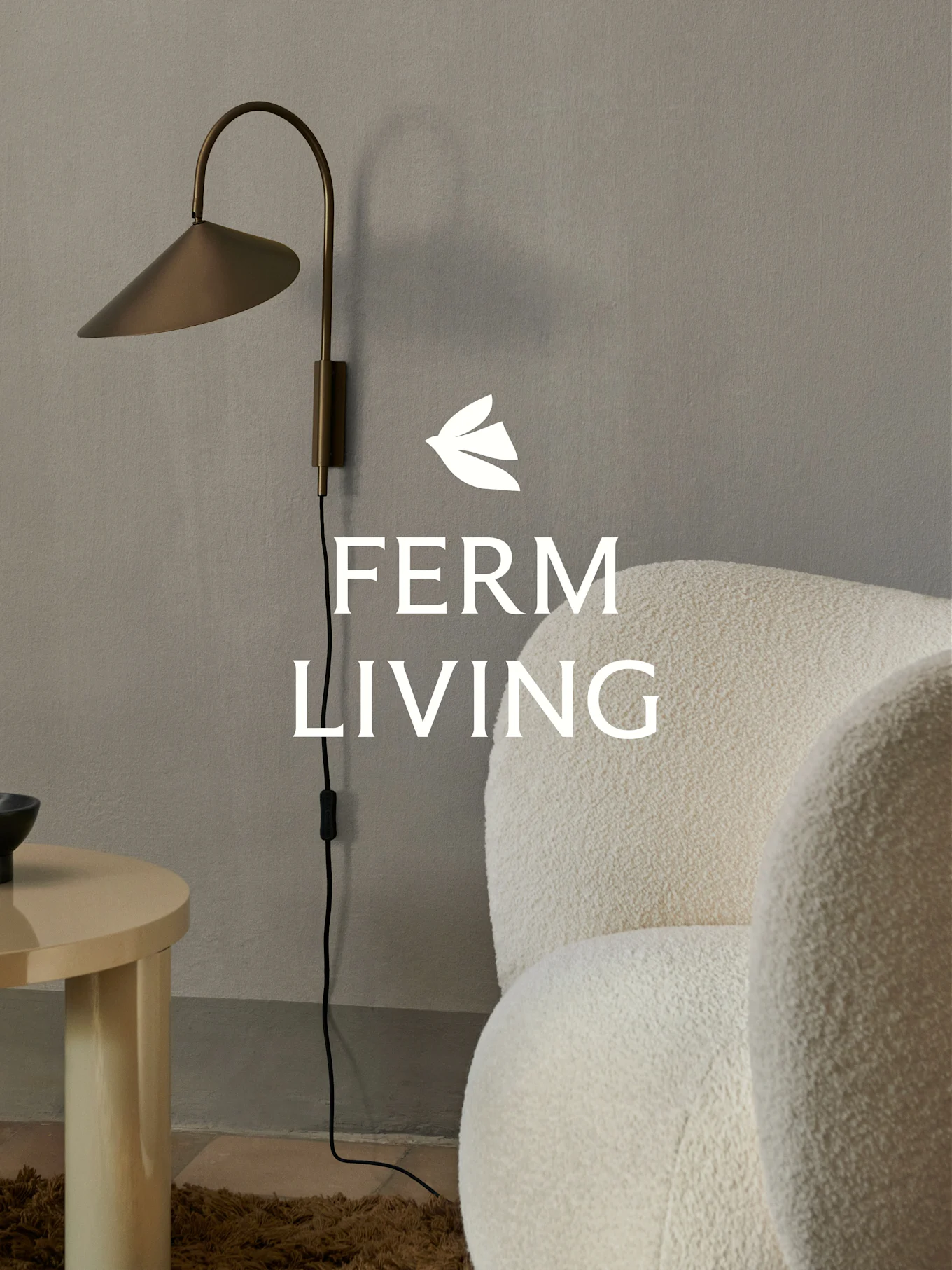 Ferm Living-logo over en messing vegglampe, en kremfarget bouclé-lenestol og et lyst sidebord i tre i minimalistisk setting.