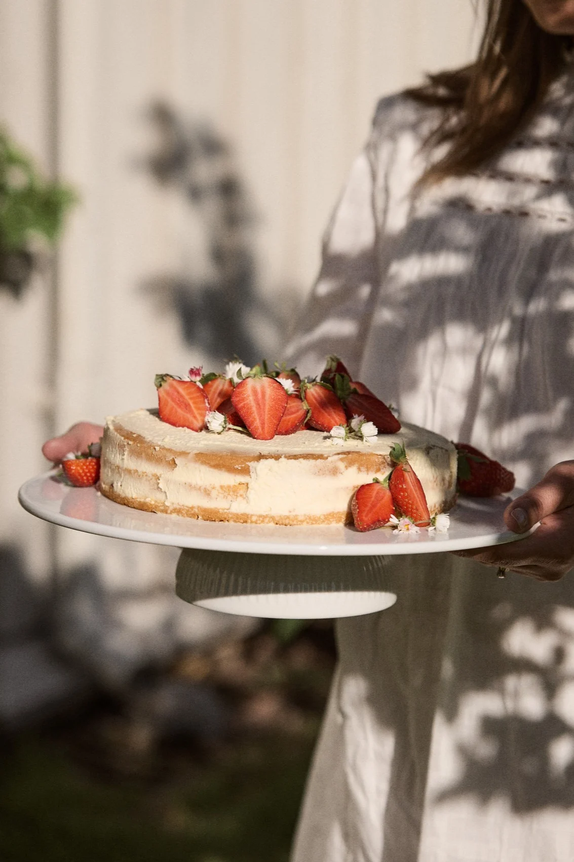 Person som holder en rund kake toppet med friske halve jordbær og små hvite blomster på et hvitt kakefat utendørs.