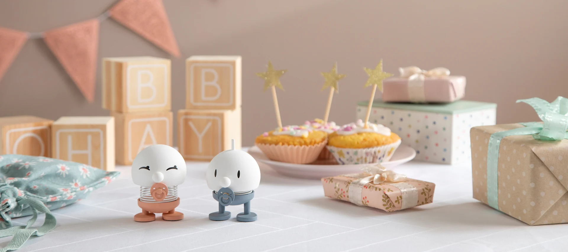 Hoptimist Baby Bimble finnes i rosa og blått og står på et bord pyntet med gaver, kranser og cupcakes.
