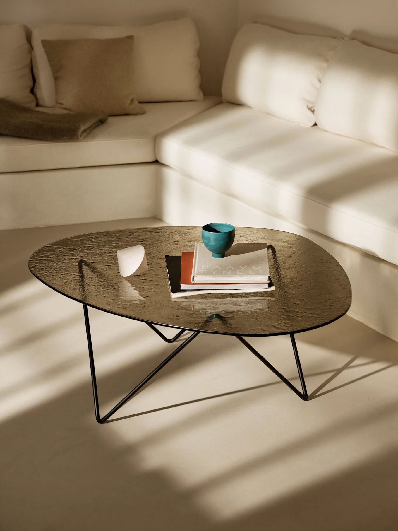 Mørkt glass salongbord med en turkis bolle, bøker og en hvit gjenstand. Lys L-formet sofa og solskinnsskygger.