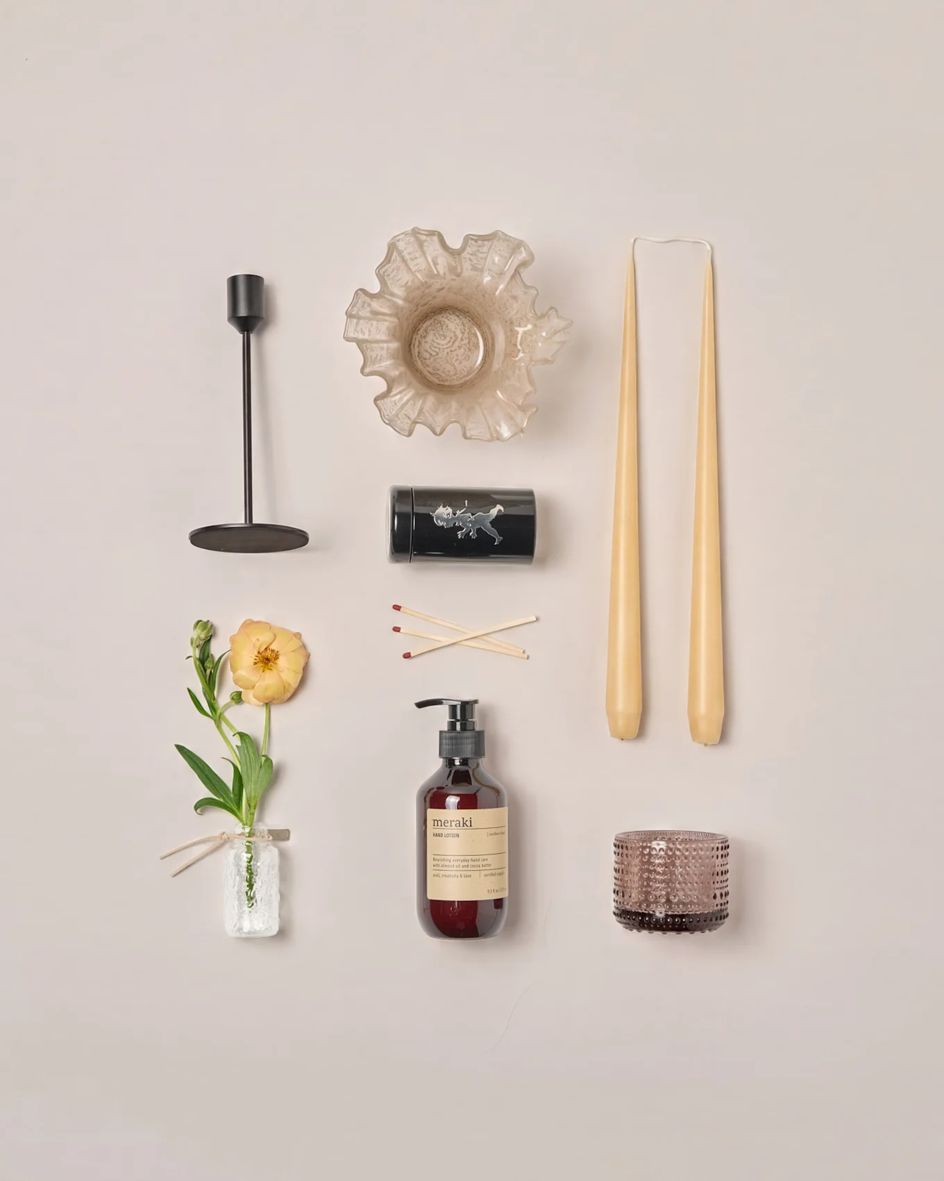 Flatt arrangement av ulike interiørartikler: lysestake, rufsete skål, koniske stearinlys, fyrstikker, lotion, telysholder og en ranunkelblomst.