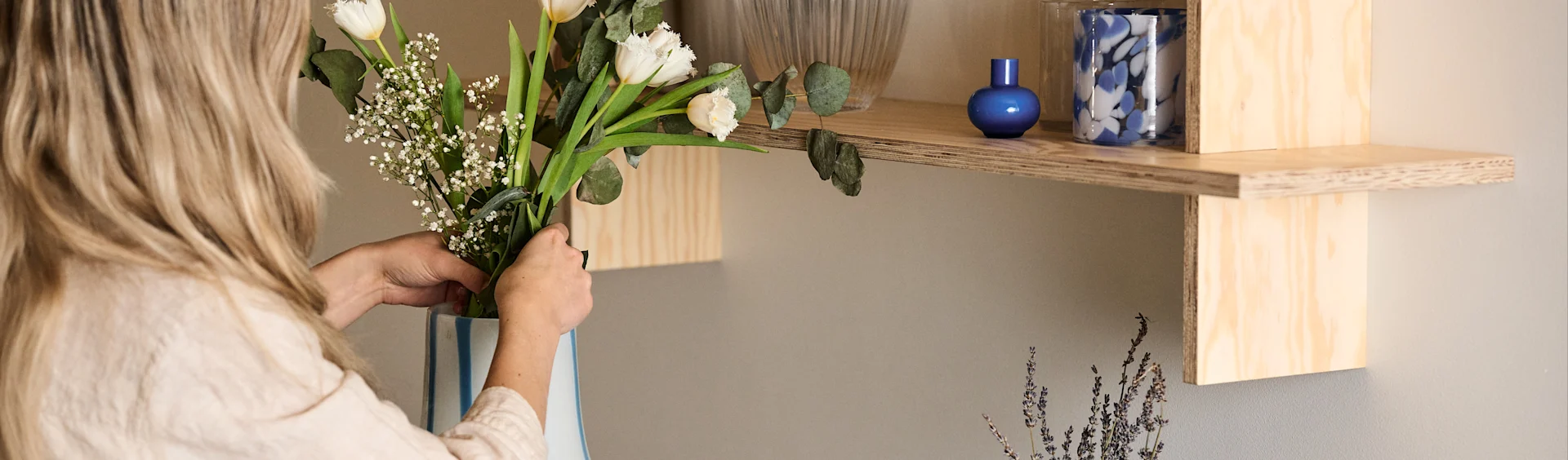 En kvinne står og justerer en bukett blomster i en Ada Stripe-vase i hvit med blå organisk formede linjer. Over vasen på en hylle står også to blå lykter.