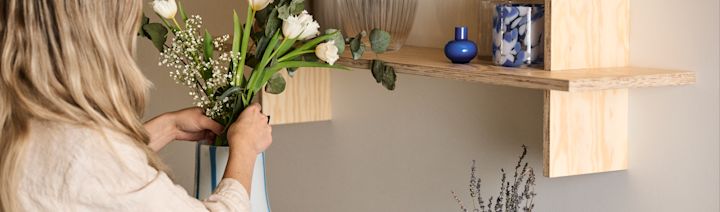 En kvinne står og justerer en bukett blomster i en Ada Stripe-vase i hvit med blå organisk formede linjer. Over vasen på en hylle står også to blå lykter.