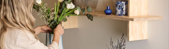 En kvinne står og justerer en bukett blomster i en Ada Stripe-vase i hvit med blå organisk formede linjer. Over vasen på en hylle står også to blå lykter.