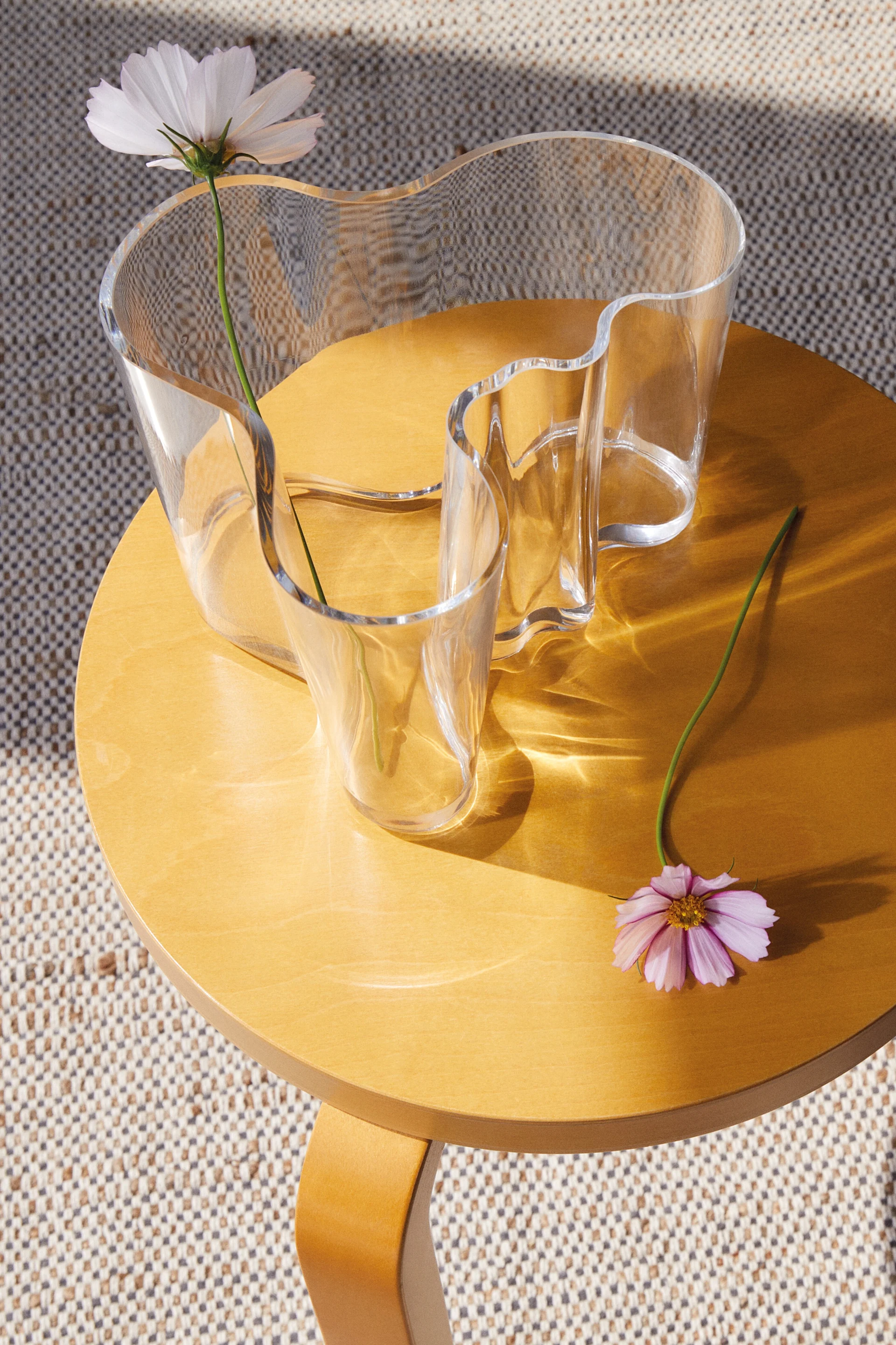 En klar, bølget glassvase med en hvit blomst på et rundt trebord, med en rosa blomst liggende ved siden av. Sollys kaster mønstre.