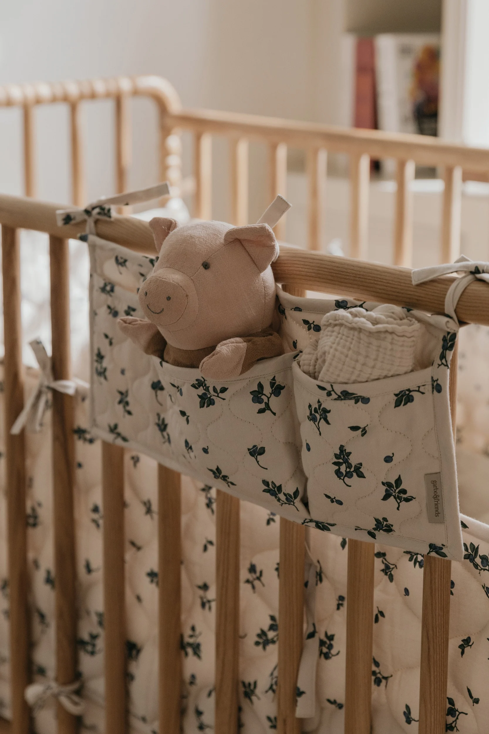 En rosa kosegris og en hvit musselinklut i en krem- og blåblomstret organizer som henger på en babyseng i tre.
