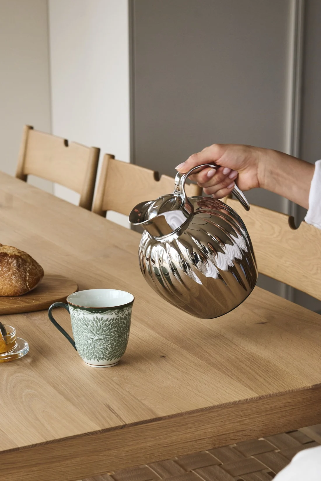 En hånd heller fra en sølvfarget riflet termokanne i et grønt mønstret krus på et trebord med brød og syltetøy.