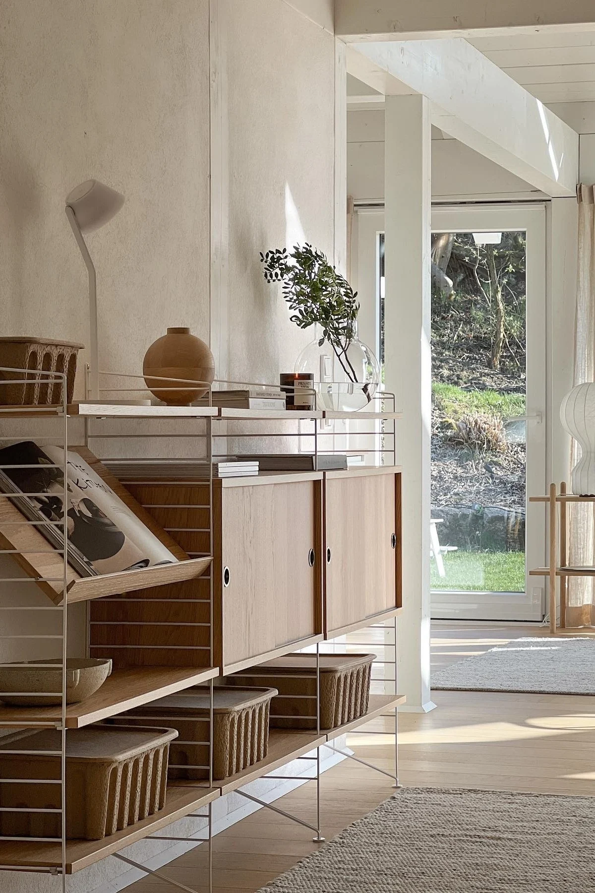 Lyst, moderne rom med hylle i lyst tre og hvite rammer, fylt med vase, bøker, grønt og kurver, med hageutsikt.