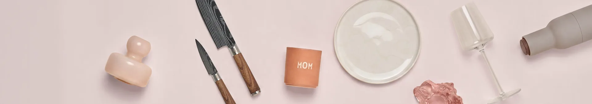Assortert kjøkkenutstyr og pyntegjenstander på rosa bakgrunn, inkludert kniver, en tallerken, et vinglass og en 'MOM'-beholder.