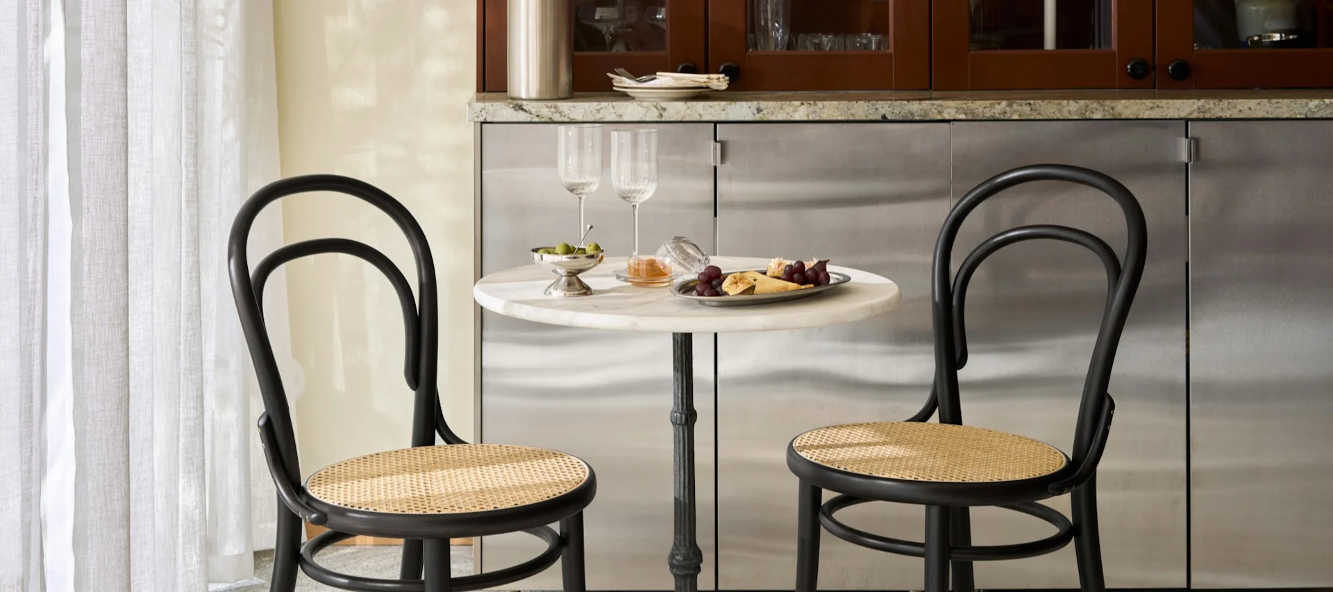 To svarte bistrostoler med rørflettede seter ved et rundt marmorbord med vinglass, oliven, druer og ost.