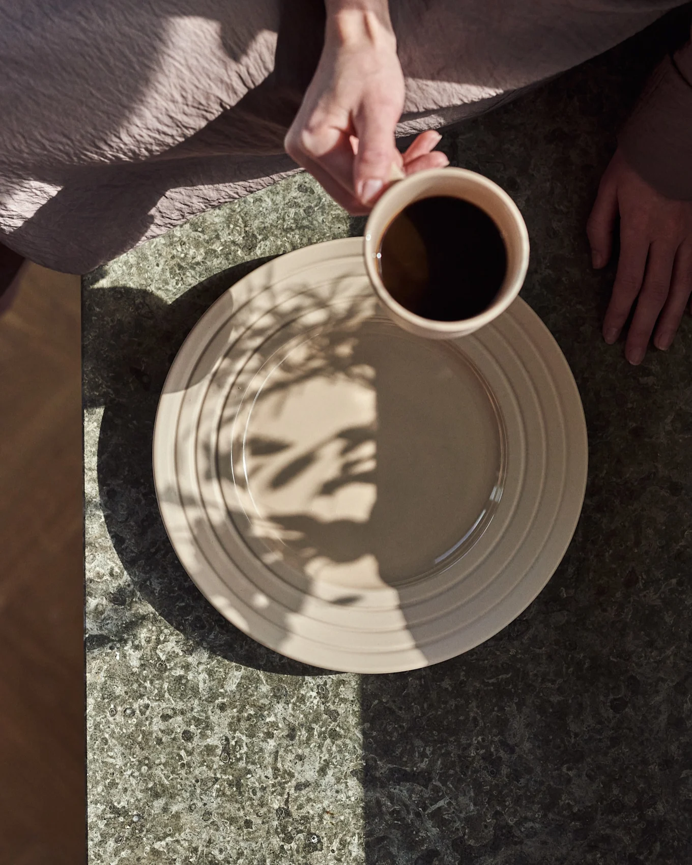 Ovenfra: hender, en holder en beige kopp med svart kaffe over en beige tallerken på et mørkt steinbord, med bladskygger.