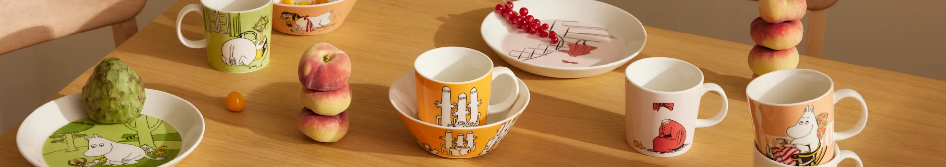 Moomin servise i ulike farger og design, med krus, skåler og tallerkener, sammen med frisk frukt på et trebord.