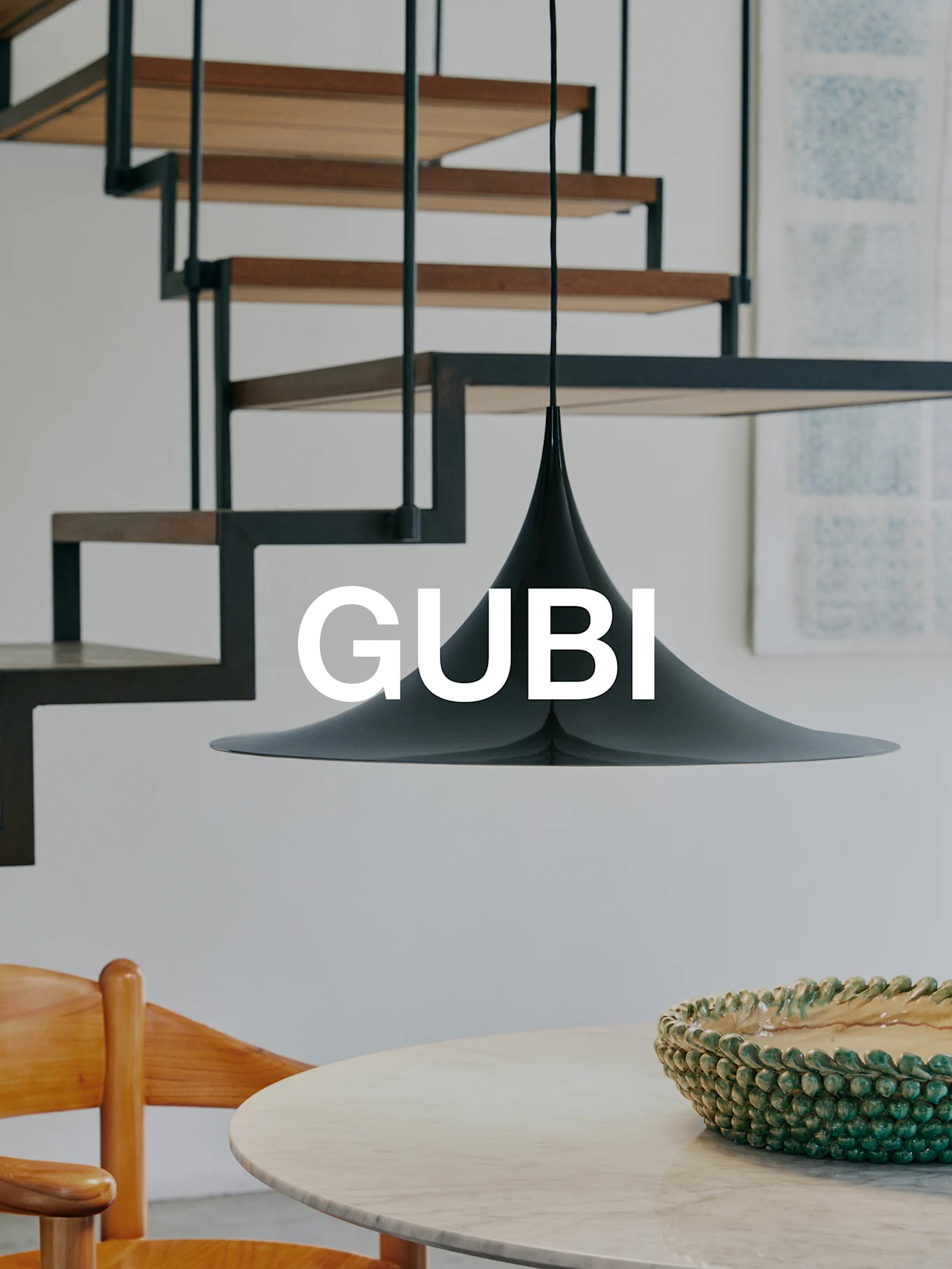 En svart konisk pendellampe over et marmorbord med en grønn skål og en trestol. En moderne trapp i bakgrunnen. Tekst 'GUBI'.