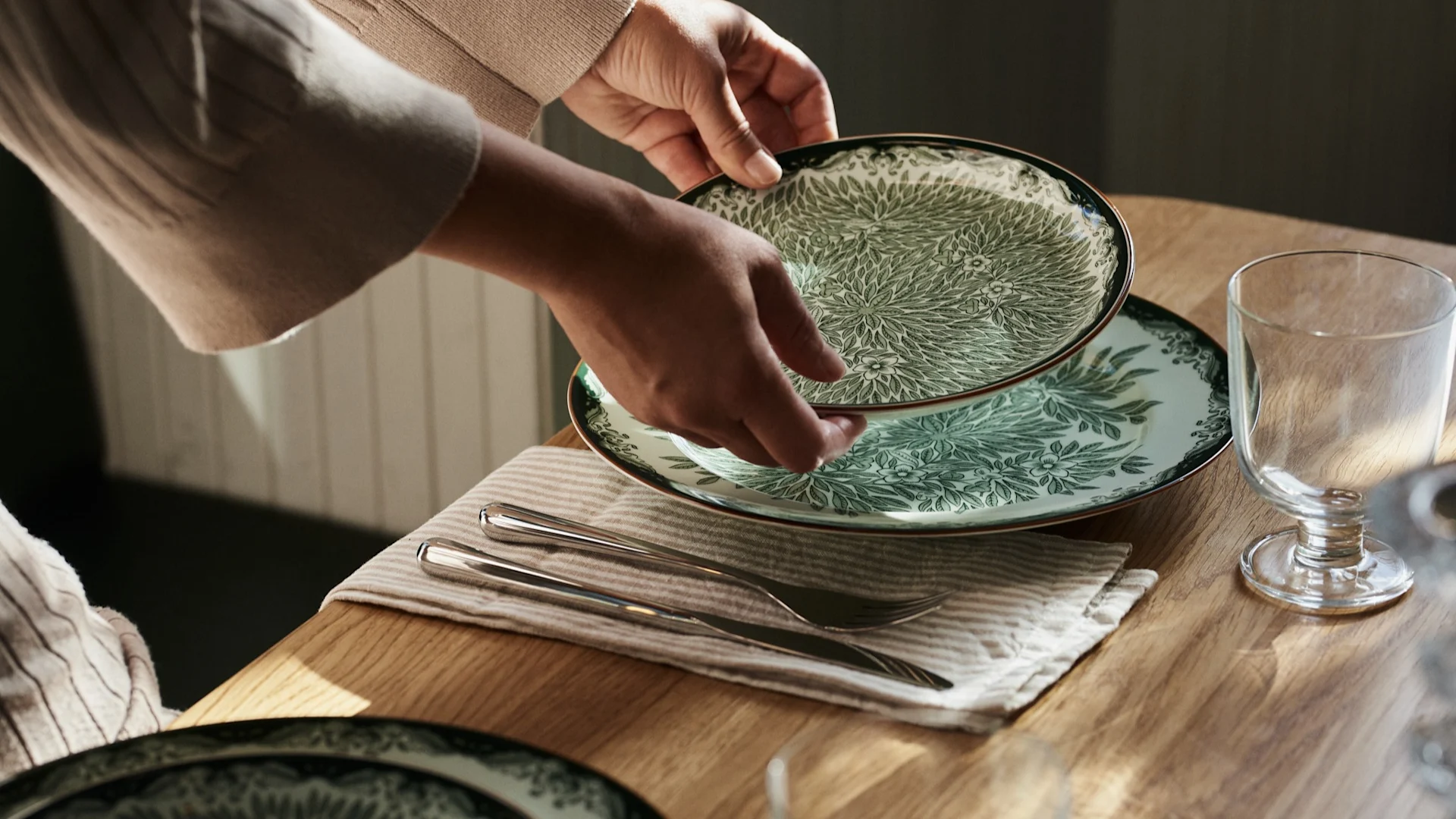 Hender plasserer en grønn- og hvitmønstret tallerken på et trebord med bestikk og et glass.