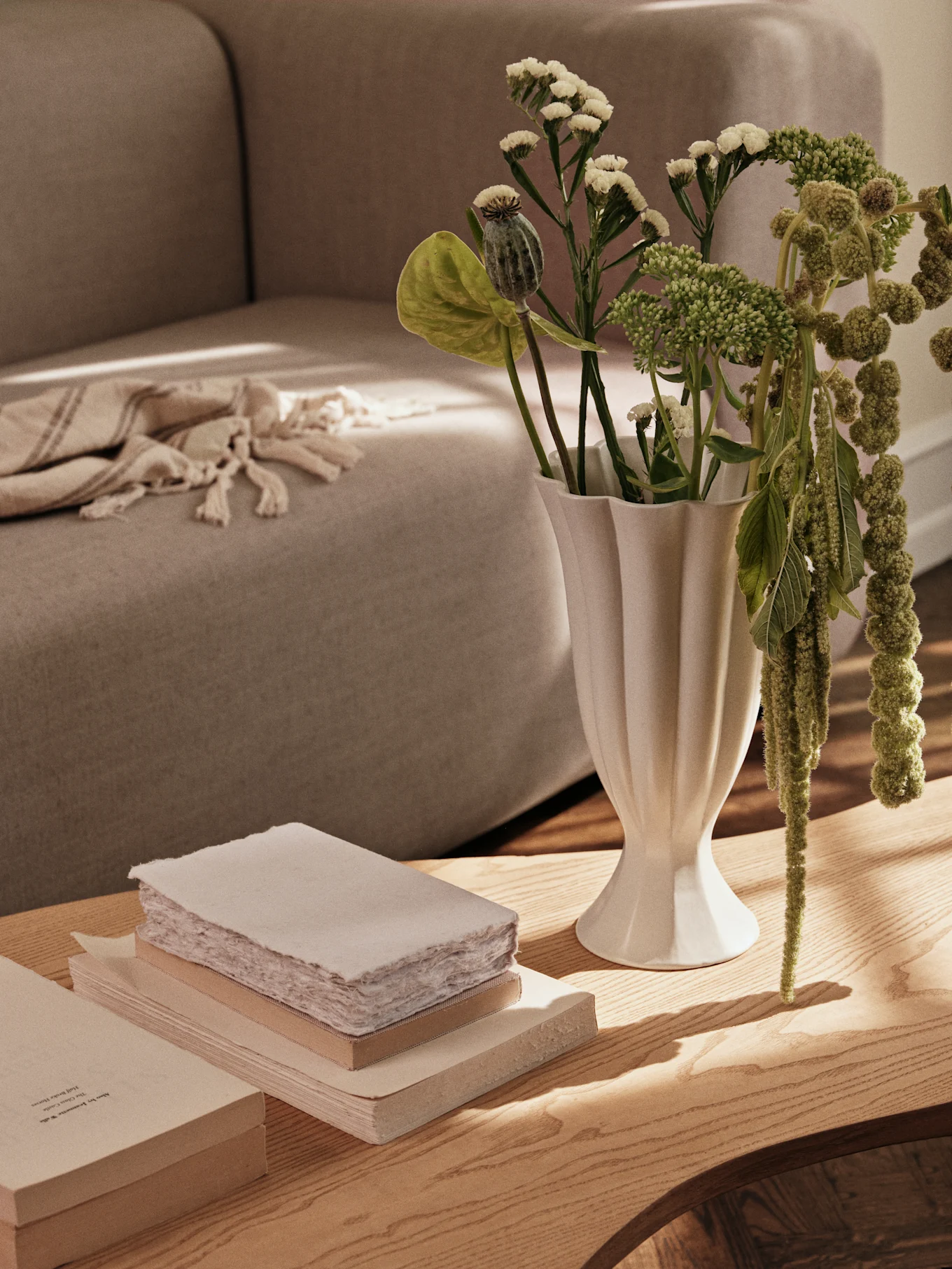 En hvit riflet vase med grønne planter og en stabel bøker på et salongbord i tre, med en sofa i bakgrunnen.