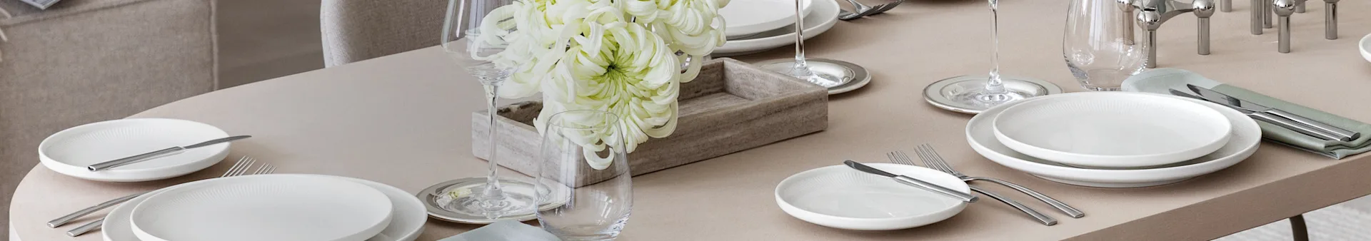 Et elegant dekket spisebord med hvite tallerkener, sølvbestikk, vin- og vannglass, og en midtdel med store hvite blomster i en trekasse.