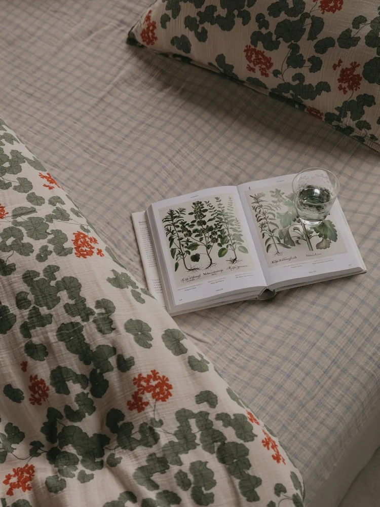En åpen botanisk bok og et glass vann på en seng med rutete dynetrekk og puter med blomstermønster.