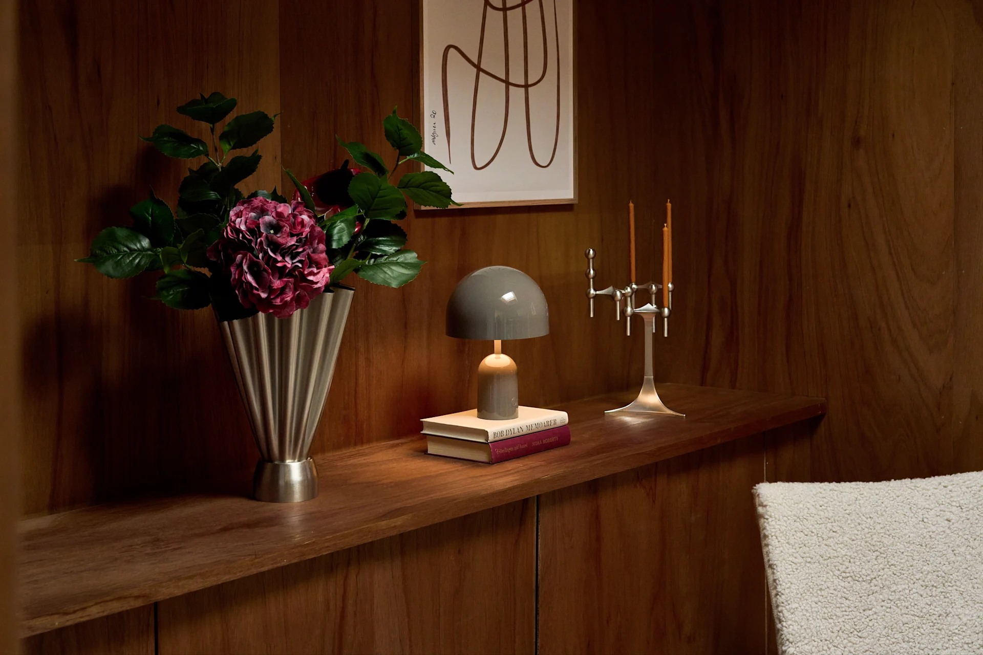 En trehylle viser en sølvfarget riflet vase med lilla blomster, en grå sopplampe på bøker og en sølv lysestake med to tente lys.