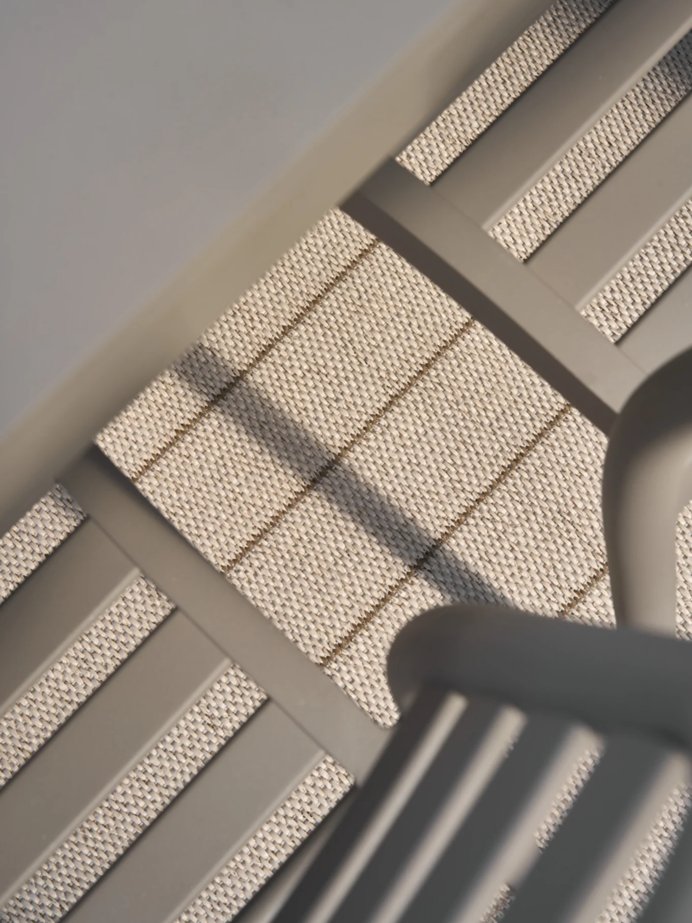 Nærbilde, vinklet visning av lyst beige vevd materiale med subtile horisontale og diagonale brune striper, innrammet av glatte grå bjelker.