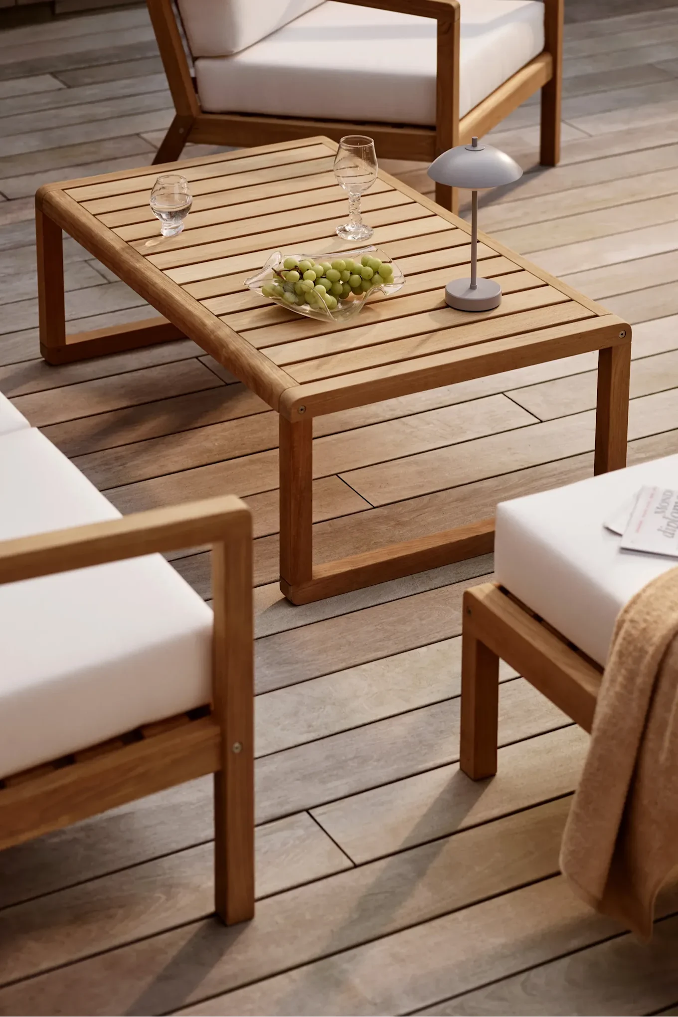 Sofabord i tre med lameller, druer, glass og Clam oppaldbar lampe, omgitt av terrassestoler i tre med hvite puter på en terrasse.