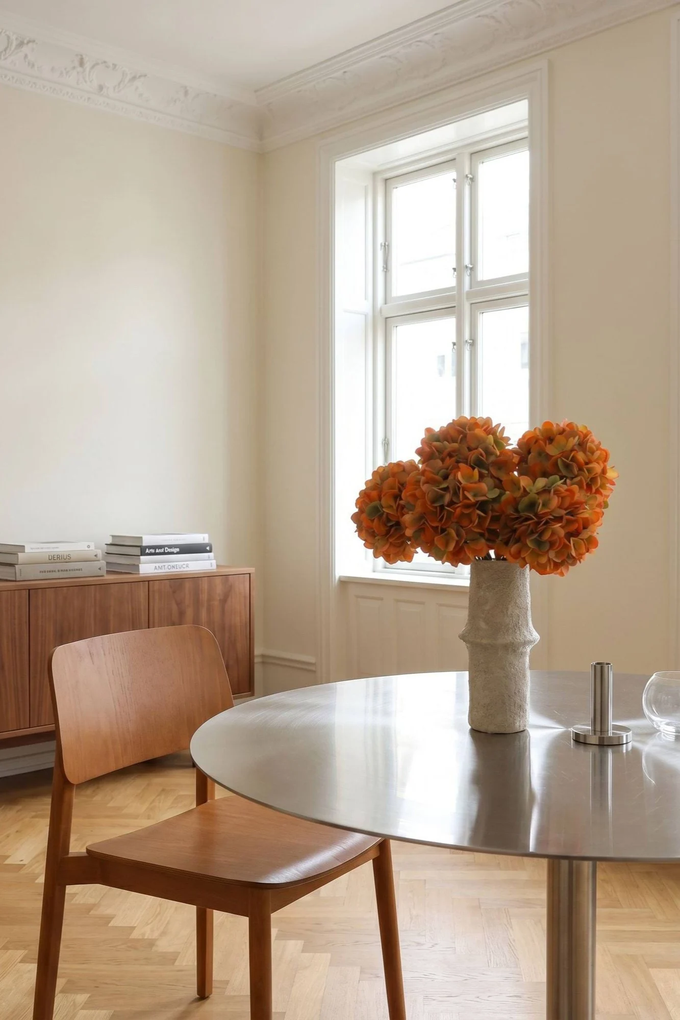 En lys spisestue med et rundt metallbord, en trestol og en vase med oransje blomster.