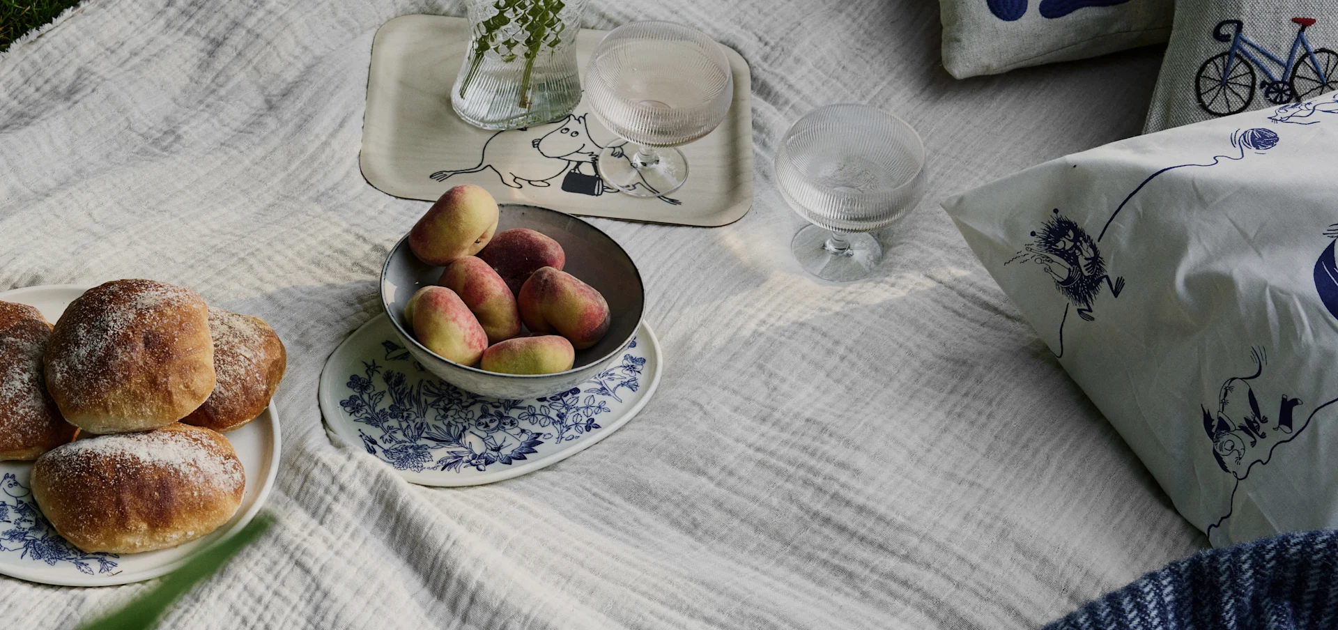 Piknikscene på et hvitt teppe med brød, fersken, drikke og puter med Moomin- og sykkelmønstre.