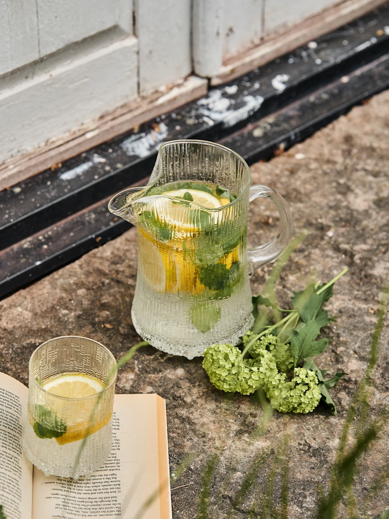 Rillet glasskanne og matchende glass med sitron- og myntevann, ved siden av en åpen bok og grønne blomster på stein.